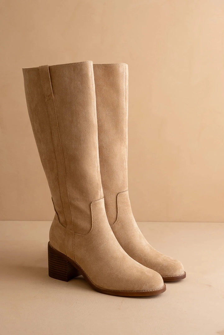 A pair of tall knee-high suede boots in a light tan/beige color, featuring a stacked block heel and a pointed toe, set against a matching beige background.