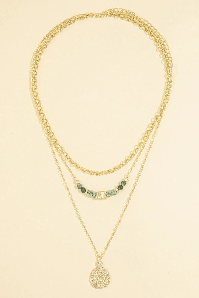 A gold layered necklace set featuring a short curb chain, a longer chain with small green and white beads, and a delicate chain with a round filigree pendant, displayed on a light beige background.