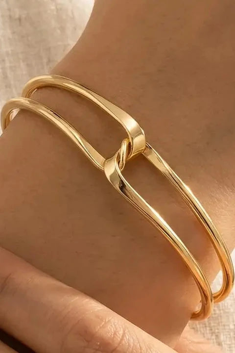 A close-up of a person's wrist wearing two slim, polished gold bangle bracelets with an interlocking twist design, set against a soft beige fabric background.